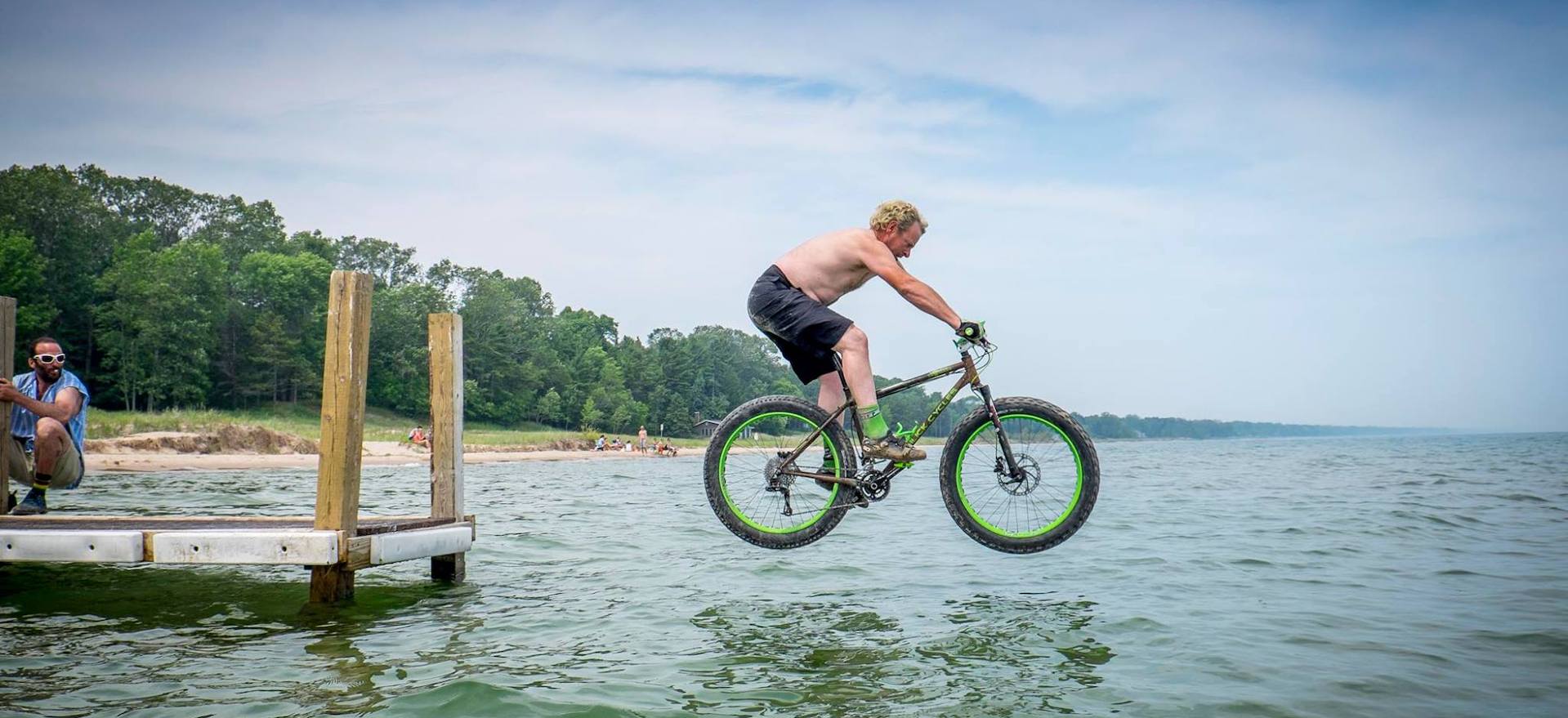 shirtless man on fat bike jumping off pier into lake