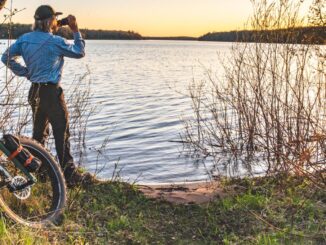 Man drinks water next to fat bike looking at sunset over lake
