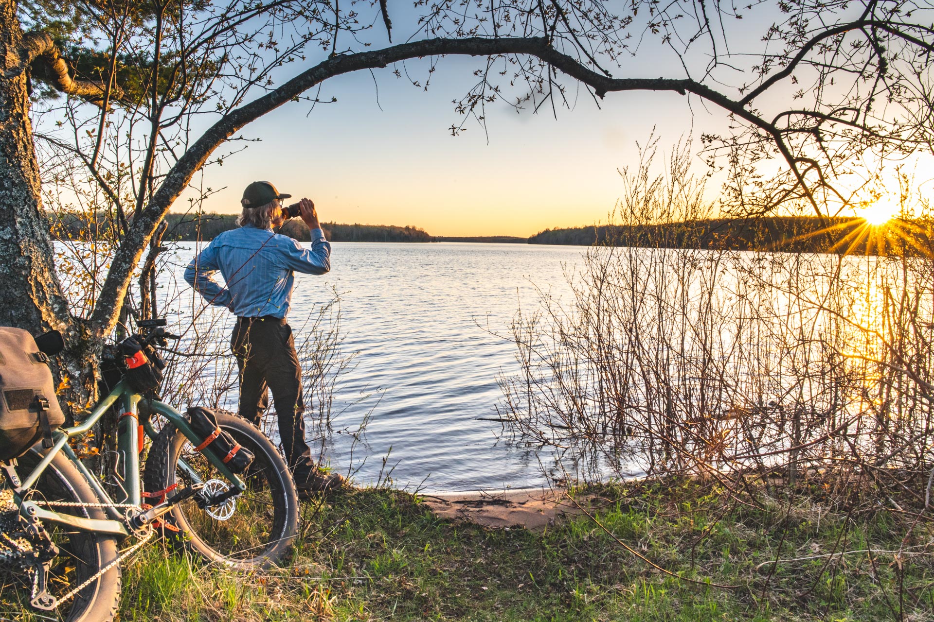 Man drinks water next to fat bike looking at sunset over lake